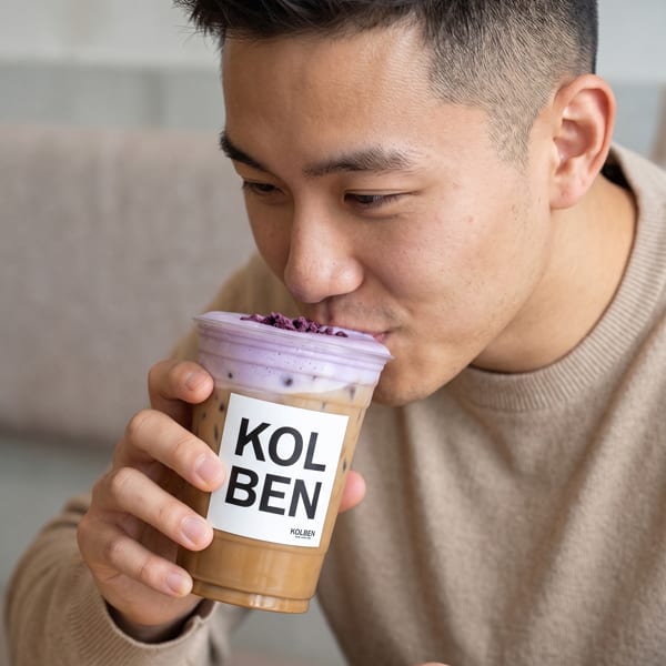 A man sips a Kolben iced coffee with purple foam and sprinkles from a clear cup.
