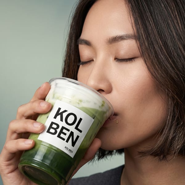 A person with closed eyes sips a green Kolben drink with a white foamy top.