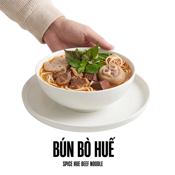 A hand holding a bowl of Bún Bò Huế, a spicy Vietnamese beef noodle soup.