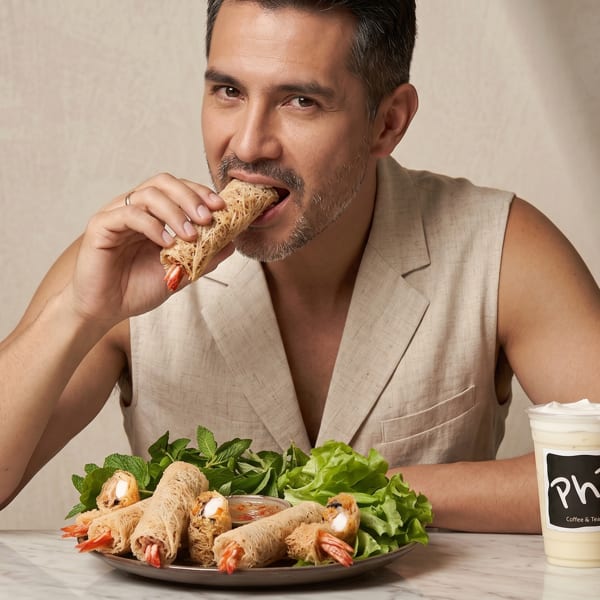 A man in a beige vest bites into a spring roll, with more rolls and Phi Coffee & Tea drinks on the table.