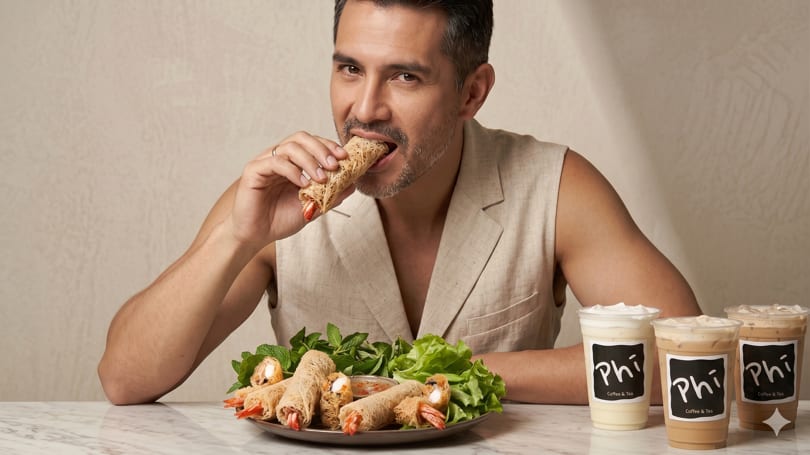 A man in a beige vest bites into a spring roll, with more rolls and Phi Coffee & Tea drinks on the table.